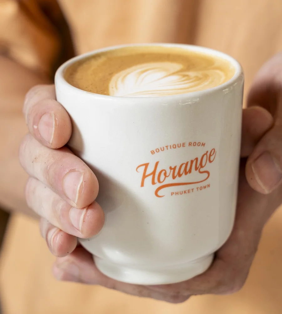 A flat white in a Horange Phuket Town cup, held in both hands at the hotel cafe